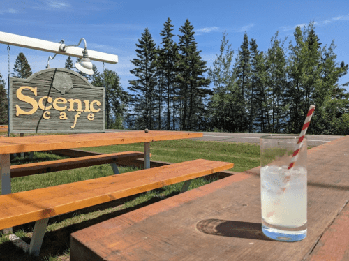 A refreshing drink with a straw sits on a wooden table near a sign for Scenic Cafe, surrounded by trees and blue skies.