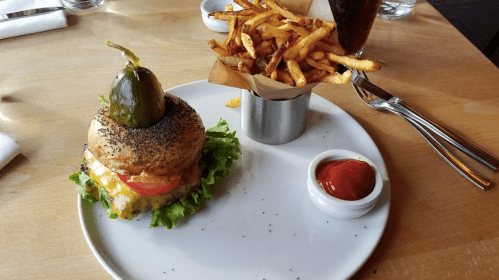 A plate with a poppy seed bun burger topped with lettuce, cheese, and a pickle, alongside a cup of fries and ketchup.