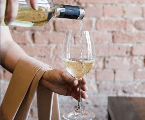 A hand pours white wine from a bottle into a glass against a brick wall background.