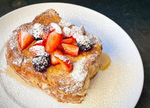 A slice of French toast topped with strawberries, blueberries, and powdered sugar, drizzled with syrup on a white plate.