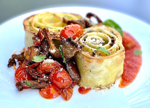 A plate featuring spiral pasta rolls with roasted tomatoes and herbs, drizzled with sauce.