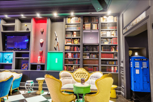 A stylish lounge with colorful seating, a bookshelf, and a blue police box in the corner.