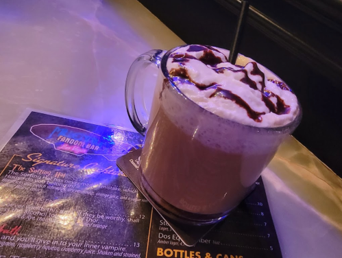 A frothy drink in a glass mug topped with whipped cream and chocolate drizzle, placed on a menu at a bar.