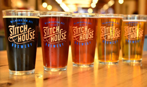 Five glasses of beer in varying colors, labeled "Stitch House Brewery," arranged on a wooden table.