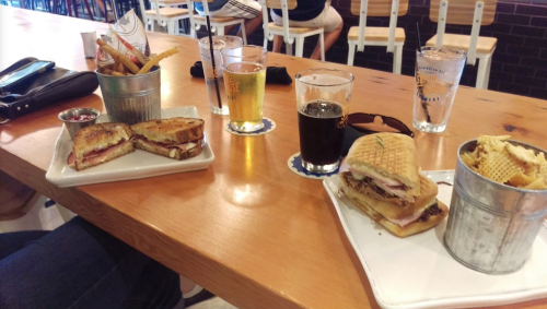 A table with two plates of sandwiches, fries in a bucket, and glasses of beer and soda.