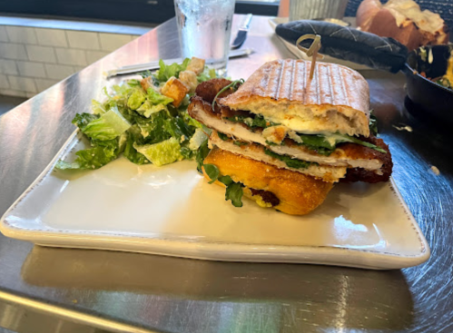 A stacked sandwich with turkey, greens, and cheese, served with a side of Caesar salad on a white plate.