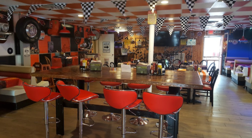 A vibrant diner interior with red stools, checkered flags, and racing-themed decor. Large wooden table in the center.