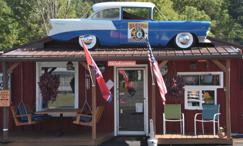 A red building with a blue vintage car on the roof, featuring flags and a welcoming sign.