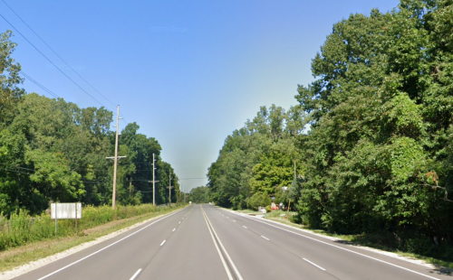 A wide, empty road flanked by lush green trees under a clear blue sky.
