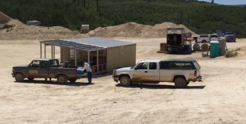 Two pickup trucks near a storage shed on a dirt site, surrounded by hills and construction materials.