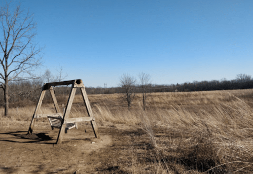 A wooden swing set stands in a barren field under a clear blue sky, surrounded by dry grass and sparse trees.