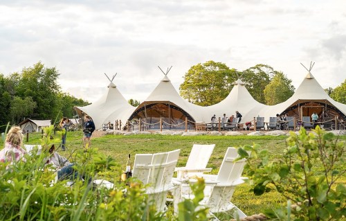 A scenic view of large white tents in a grassy area, with people mingling and trees in the background.