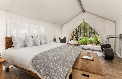 Cozy glamping tent interior with a large bed, wooden furniture, and a view of the forest through an open entrance.