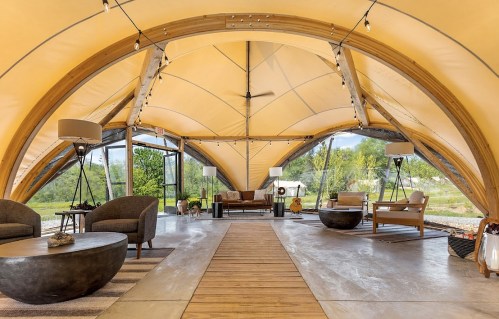 Interior of a modern tent with wooden beams, cozy seating, and large windows overlooking greenery.