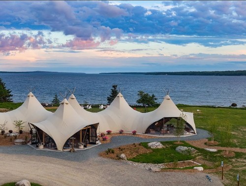 A scenic view of three large, white tents by a calm lake, surrounded by greenery and a colorful sky at sunset.