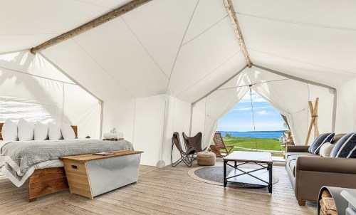 Cozy glamping tent interior with a bed, seating area, and a view of the ocean through an open entrance.