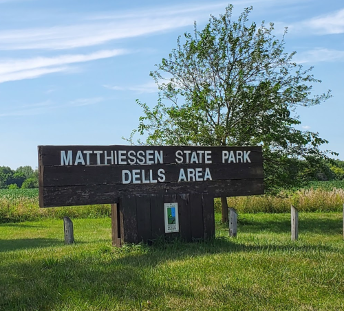 Sign for Matthiessen State Park, Dells Area, surrounded by green grass and trees under a blue sky.
