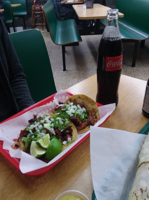 A tray with two tacos topped with onions and cilantro, lime wedges, and a bottle of Coca-Cola on a table.