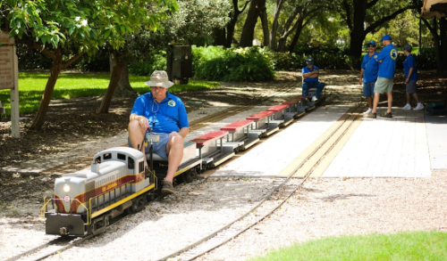A man in a hat rides a miniature train, while others in blue shirts work nearby in a park setting.