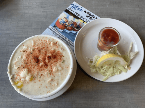 A bowl of creamy seafood chowder topped with seasoning, accompanied by a shot of cocktail sauce and a lemon wedge on a plate.