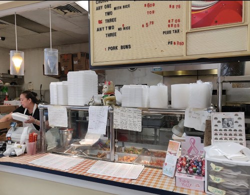 A busy takeout counter with a staff member, food containers, and a menu board displaying prices and options.