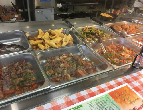 A display of various dishes in metal trays, including fried dumplings, stir-fried vegetables, and meat options.