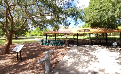 A park pavilion with a metal roof, surrounded by trees and a grassy area, featuring picnic tables and a fire hydrant nearby.