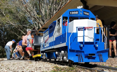 A miniature train with a blue and white design, parked near a group of people sitting on benches under a shelter.