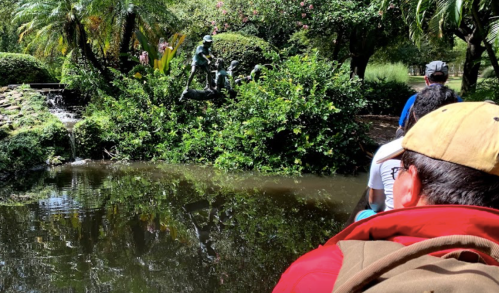 A serene pond surrounded by lush greenery, with a statue and people enjoying the view from a boat.