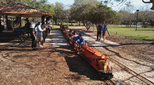 A miniature train with passengers rides along a track in a park, surrounded by trees and people enjoying the day.