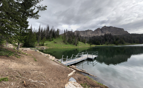 A serene lake surrounded by green hills and rocky cliffs, with a wooden dock extending into the water.