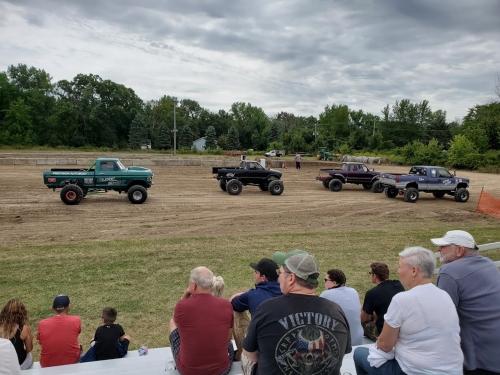 Four trucks lined up on a dirt track, with spectators seated in the foreground, enjoying a truck event.