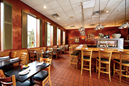Cozy restaurant interior with wooden tables, chairs, and warm lighting, featuring large windows and a bar area.