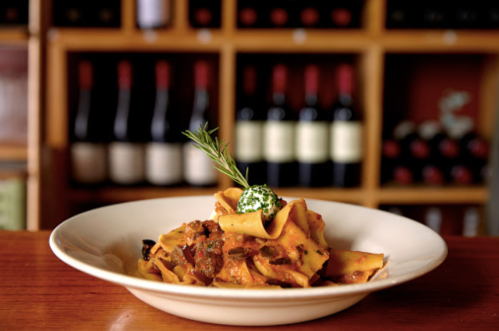A plate of pappardelle pasta topped with sauce, herbs, and a dollop of cheese, with wine bottles in the background.