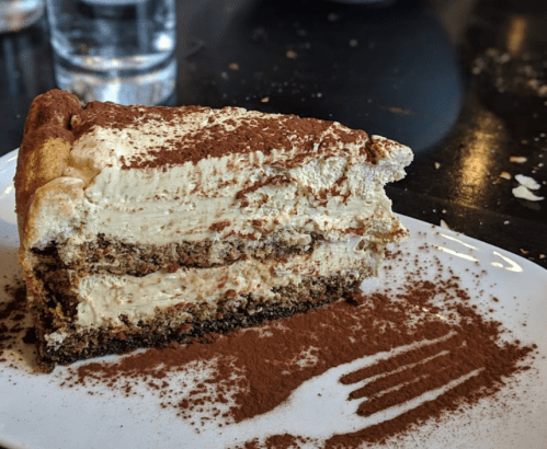 A slice of creamy tiramisu cake on a white plate, dusted with cocoa powder, with a fork resting beside it.
