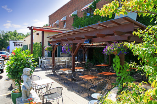 Outdoor dining area with wooden pergola, tables, and hanging flowers, surrounded by greenery and a brick building.