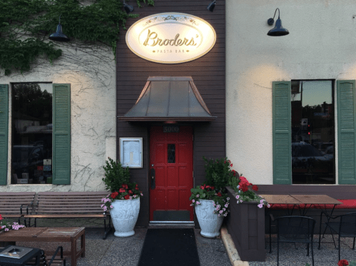 Exterior of Broder's Pasta Bar, featuring a red door, green shutters, and flower pots, with outdoor seating visible.