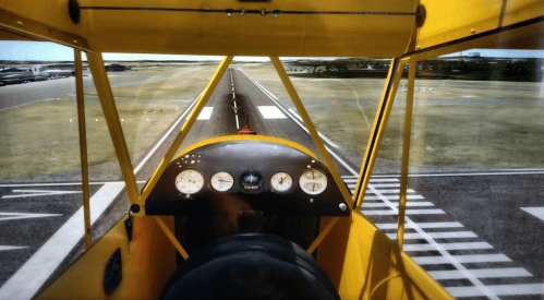 Cockpit view of a yellow biplane on a runway, with gauges visible and a clear landscape ahead.