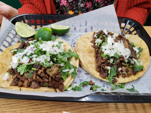 Two tacos filled with seasoned meat, topped with onions and cilantro, served with lime wedges on a black tray.