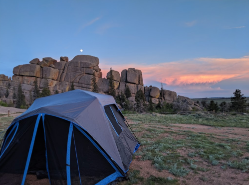 A tent in a grassy area with large rock formations and a colorful sunset in the background.