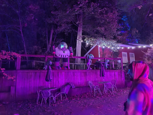 Colorful alien figures stand on a wooden railing in a dimly lit outdoor area with purple lighting and empty chairs.