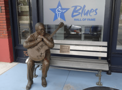 A bronze statue of a musician with a guitar sits on a bench outside the Blues Hall of Fame.