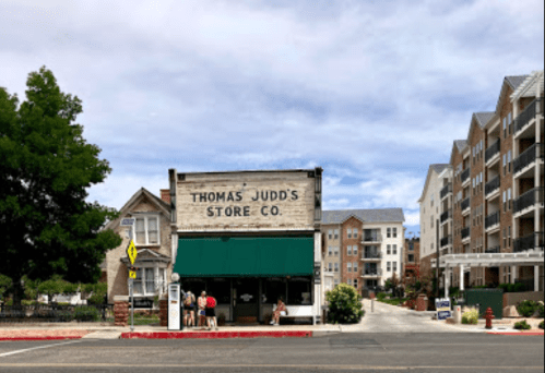 Historic storefront of Thomas Judd Store Co. with modern buildings nearby under a cloudy sky.