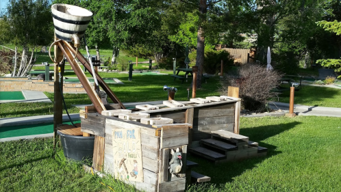 A wooden mini-golf hole featuring a water feature and a sloped ramp in a grassy park setting.