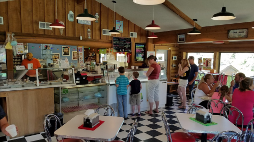 A busy ice cream shop with families ordering at the counter and enjoying treats at tables. Wood paneling and checkered floor.