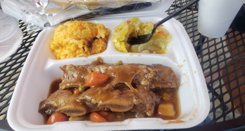 A takeout container with beef ribs in gravy, yellow rice, and a side of macaroni salad on a table.