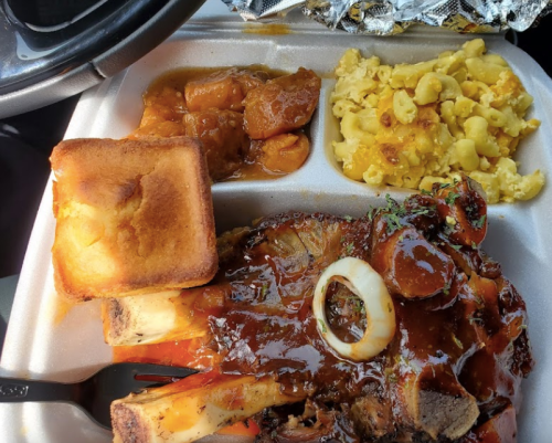 A takeout container with BBQ ribs, macaroni and cheese, sweet potatoes, and cornbread.