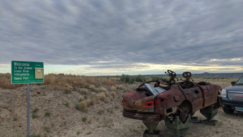 A rusty car on a stand near a sign welcoming visitors to the Greater Green River Intergalactic Space Port.