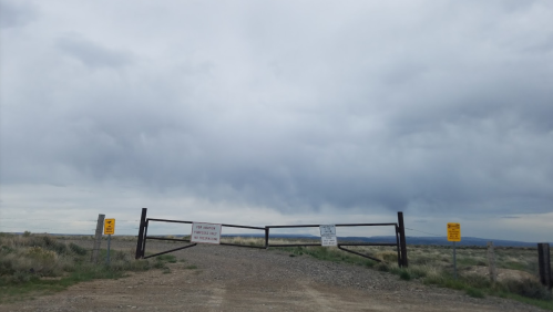 A gravel road leads to a closed gate with warning signs under a cloudy sky.