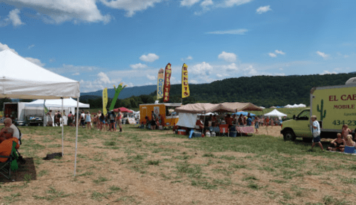 A vibrant outdoor festival scene with food stalls, tents, and people enjoying the day against a mountain backdrop.